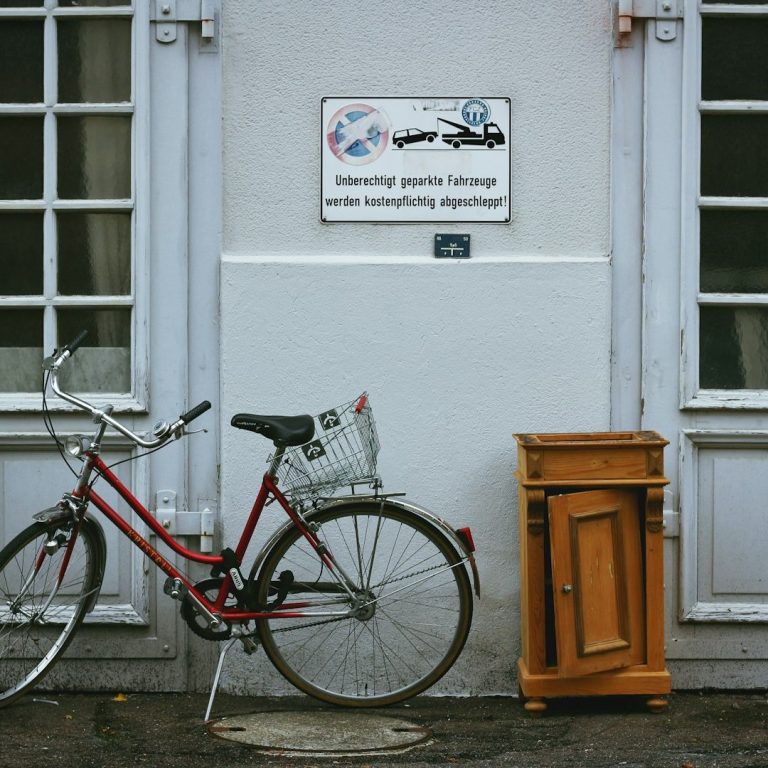 Rotes Fahrrad neben einem braunen Holzschrank an einer Wand mit Fenstern.